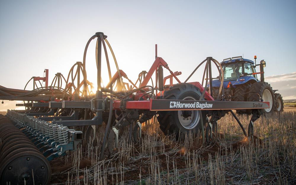 A Horwood Bagshaw machine being towed by a New Holland tractor through a field of stubble at sunset
